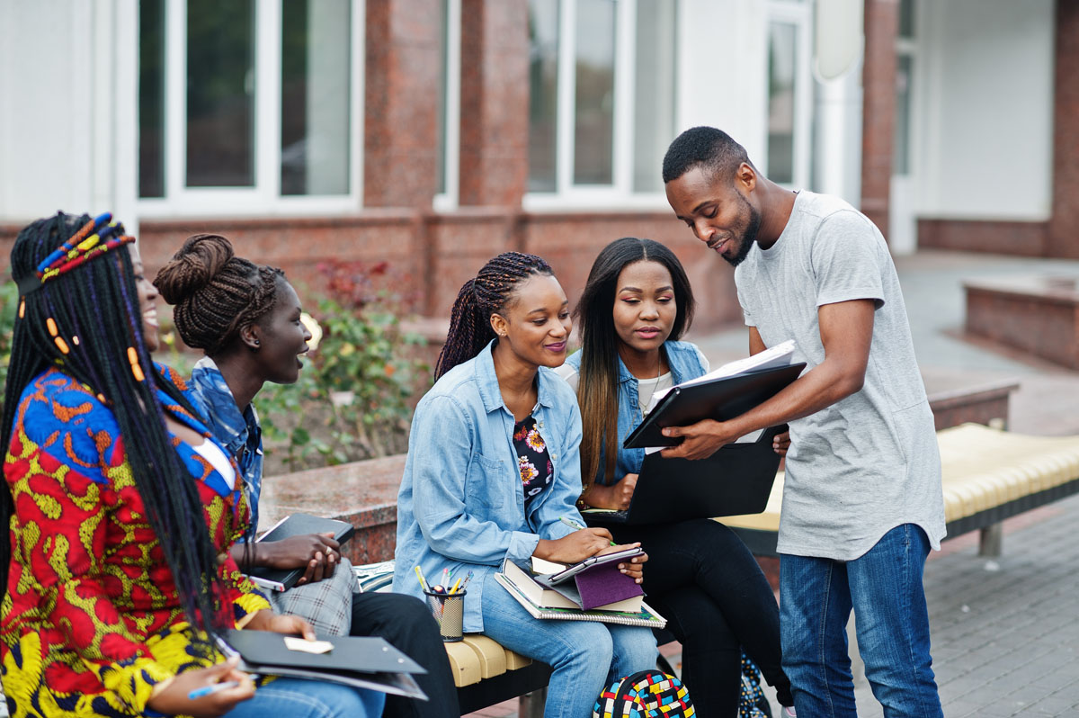 Group of five african college students spending time together on campus at university yard. Black afro friends studying at bench with school items, laptops notebooks.