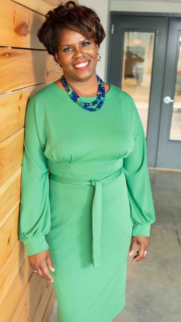 Michelle Bolden, Call for Caring. Wearing a green dress, standing up and leaning on a wall.
